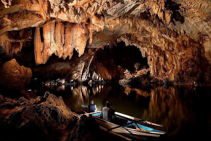 Two people in a boat tour navigate a dimly lit cave with stalactites, surrounded by calm water reflecting the cave walls.