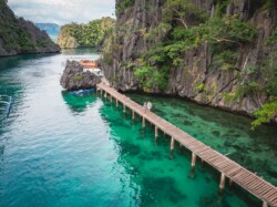 A wooden walkway extends over the clear turquoise waters of Coron, surrounded by rocky cliffs and lush greenery. Two people walk hand in hand along the pathway, with boats visible on the water. The sky above Palawan is partly cloudy, adding a dreamlike charm to the scene.