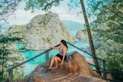 A person sits on a rock ledge in Palawan, soaking in the scenic view of turquoise waters and lush, rocky islands of Coron. The scene is framed by trees, with wooden railings and a small pathway visible in the foreground.