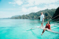 A man and woman enjoy a sunny day on a boat in the turquoise waters of Palawan. The man stands, wearing a light shirt and shorts, while the woman relaxes, dressed in red, against a backdrop of Coron’s lush green mountains under a clear blue sky.