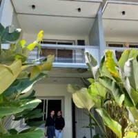 Bright hotel balcony with white walls and black railings, lush green plants in the foreground, sunny.