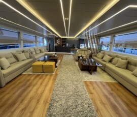 A spacious yacht interior features beige sofas, wooden tables, soft carpet, and panoramic windows offering scenic views, ideal for travel comfort. Comfortable beige sofas, wooden tables, and natural lighting create a cozy yacht lounge. Perfect for relaxing during island travel.