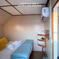 Interior of a cruise cabin featuring a double bed, pillows, and essential amenities for a comfortable stay. Cozy double bed in Manta Queen 3 cruise cabin with amenities.